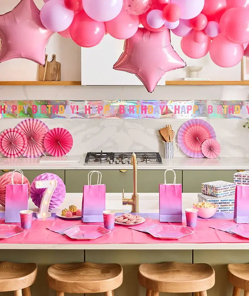 Pink birthday party table with balloon garland, star balloons, a lit number 7 decoration, gift bags, party tableware, and birthday decorations.